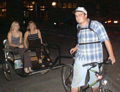 Photo of James M. Branum driving a pedicab (a mountain bike pulling a trailer), with two passengers on board.