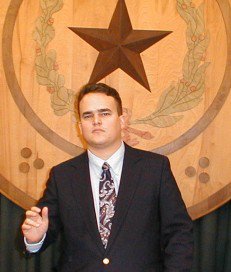 picture of James M. Branum, Feb. 1, 2000 at the Texas State Capitol in AustinThe picture is of a young man in his 20's with brown hair. He is wearing a suit and is standing in front of a wooden plaque of the seal of the state of Texas