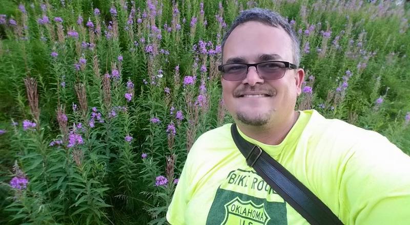 picture of a middle aged man wearing a fluorescent yellow shirt, in front of bunch of fireweed growing in Anchorage, Alaska