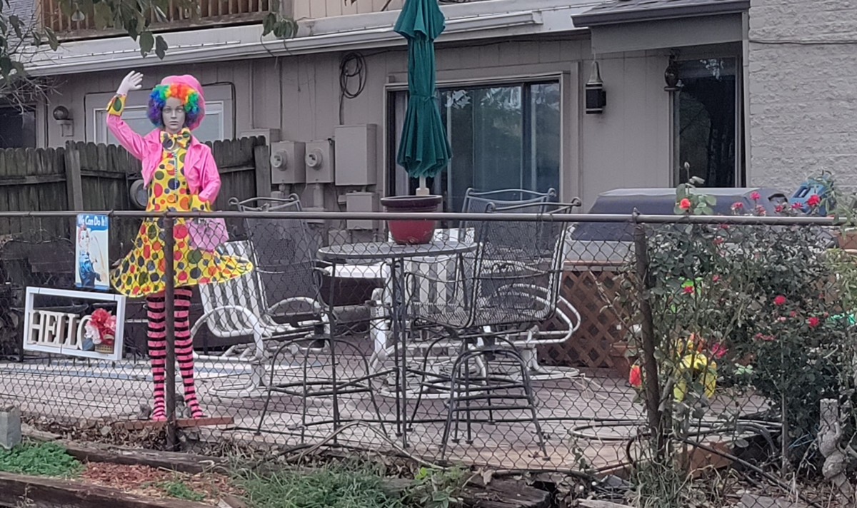 picture of a back patio with a maniquin standing and waving, with rainbow colored hair, and a pretty dress. There is sign that says Hello and there is a rosie the riveter poster nearby.